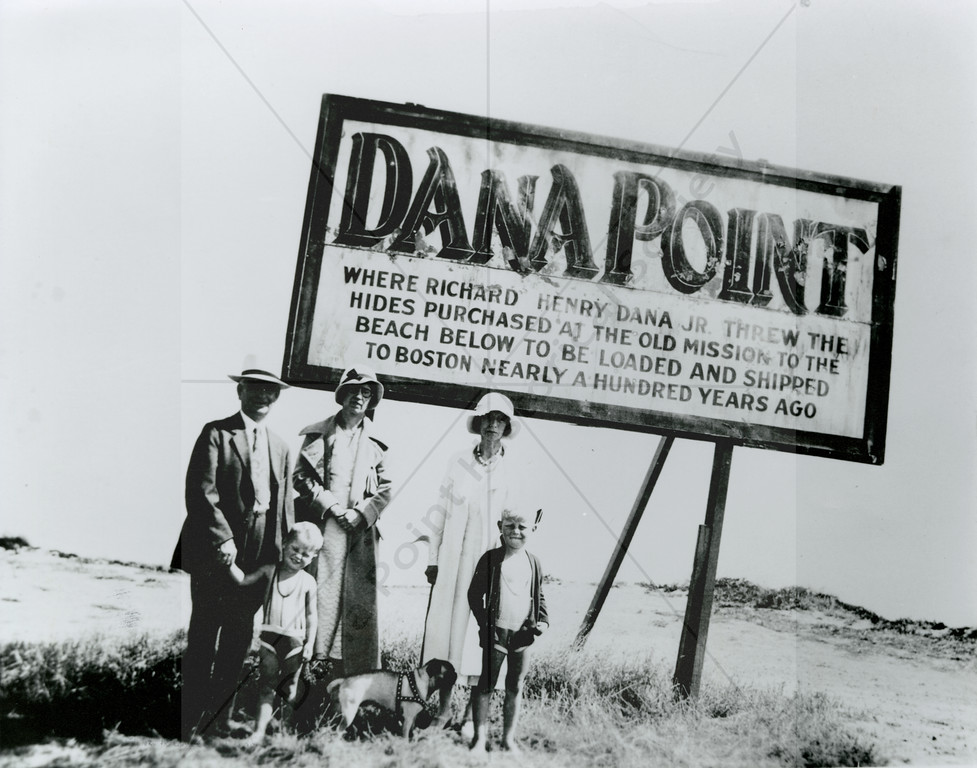 Historic black-and-white photograph of three adults, two children and a small dog standing beneath a large sign reading “Dana Point — Where Richard Henry Dana Jr. threw the hides purchased at the old mission to the beach below to be loaded and shipped to Boston nearly a hundred years ago”