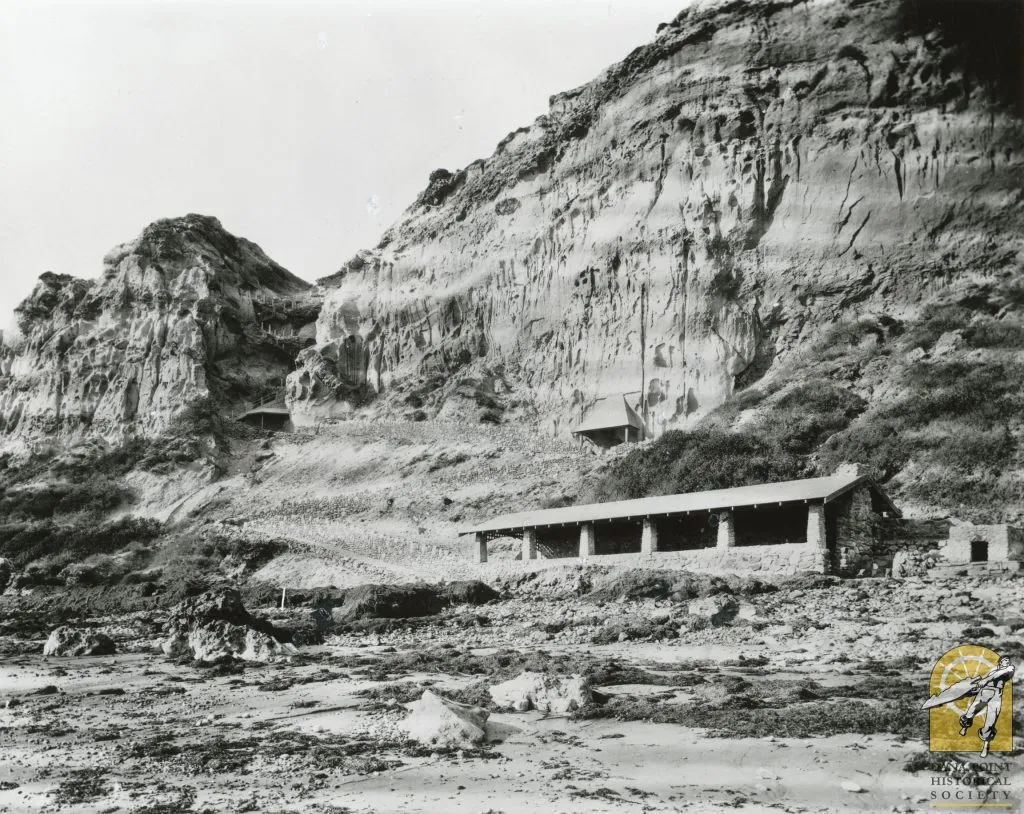 Black-and-white photo of a low building at the foot of eroded coastal bluffs